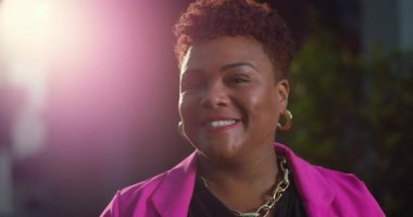 Afro-Amerikan kadın, kısa doğal saçlı, kameraya nazikçe gülümseyen, altın takılar içinde rahat ve kendine güvenen, pembe ceketli.