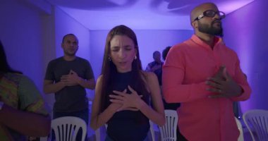 İbadet sırasında gözleri kapalı ve elleri göğsünde duran genç bir kadın. Etrafı dua eden cemaatin üyeleriyle çevrili.