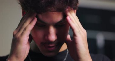 Latin kökenli genç bir adam parmaklarını şakaklara bastırıyor, gözler aşağıda, odaklanmış ifade modern bir iç mekanda zihinsel baskı veya yorgunluk olduğunu gösteriyor.