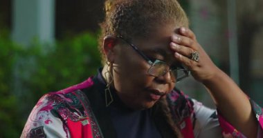 Afro-Amerikan kadın başını eğiyor ve gözle görülür bir stres anında tapınaklara ellerini bastırıyor, açık havada, mahcup bir ifadeyle ve gözleri kapalı duruyor.