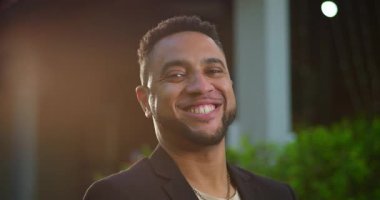 Akşamları gülümseyen Afro-Amerikalı adam kameraya bakıyor, sıcak tonlarda ve yumuşak kentsel arka planda ince mercek parıldıyor.