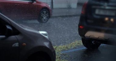 Park halindeki arabaların arasına sağanak yağmur yağıyor. Sarı yapraklar mavi park alanı boyunca birikiyor ve kırmızı araç arkadan geçiyor.