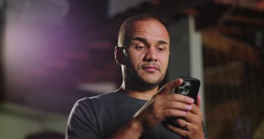 Afrika kökenli Latin kökenli bir adam dışarıda akıllı telefon kullanıyor. Akşamları iki eliyle telefonu tutarken internet içeriğine tepki veriyor.