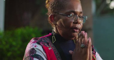 Olgun Afrikalı Amerikalı kadın, elleri kapalı ve gözleri kapalı, duacı bir şekilde ayakta duruyor. Doğada ruhani yansımanın huzurlu bir anı.