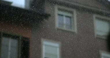 Soft-focus rainfall in front of urban residential buildings with visible window frames and faint storm patterns in slow-motion detail