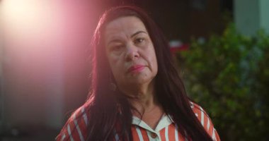 Uzun siyah saçlı ve çizgili bluzlu Latin kadın kameraya ciddi bir ifadeyle bakıyor. Açık havada hafif merceklerle dalgın dalgın bakıyor.