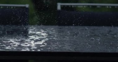 Yavaş çekimde cam veya metal masa yüzeyinden yansıyan yağmur damlaları kabarcıklar ve su sıçratan taç, koyu renk sandalyeler ve arkadaki yapraklarla keskin bir yakın plan çekimde yakalanır.