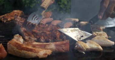 Izgara kaburgalar, tavuk kanatları ve sosisler dumanlı barbeküde pişerken iki metal spatula ile et çeviriyorlar, yüksek hızlı kamerayla süper yavaş çekimde çekiliyorlar.