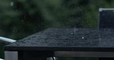 Yüksek hızlı kamerayla çekilen süper yavaş yağmur, metal masa köşesine çarpıyor, damlalar havada uçuşuyor, arka bahçede bulanık yeşil ağaçlar var.