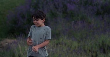 Çocuk lavanta tarlasına düşen yağmura tepki veriyor, neşeli bir gülümseme ve ıslak gömlekle eğiliyor, yavaş çekimde açan mor ve yeşil bitkilerle çevrili.