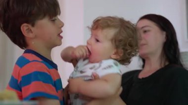 Daha büyük bir çocuk kucağında bebek taşıyarak anne gözetiminde görünür çaba ve özen gösteriyor. Kapalı alanda özgün bir kardeşlik bağı ve aile içi etkileşim anı yaşıyor..