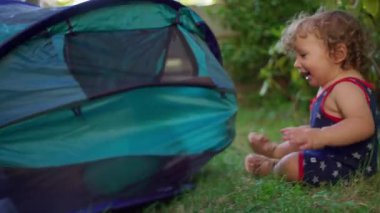 Gülen bebek, güneşli arka bahçedeki çadıra doğru emeklerken ağabeyi içeride saklanıp kumaşı tıkırdatıyor, neşeli ve kaygısız bir kardeş eğlencesi anı yaratıyor.