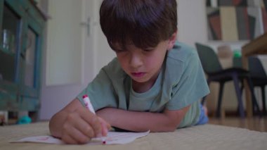 Oğlan, dirsekleri üzerinde dinlenirken, evde yerde yatarken, kırmızı kalem kullanarak, sessiz yaratıcı aktivitelere dalarken kağıt çizer.