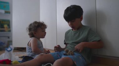 Büyük kardeş anahtarlık pelüş oyuncakla oynuyor. Küçük kardeş merakla izliyor. Her ikisi de arka planda kitap ve oyuncaklarla dolu, rahat bir odada, ahşap zeminde oturuyor.