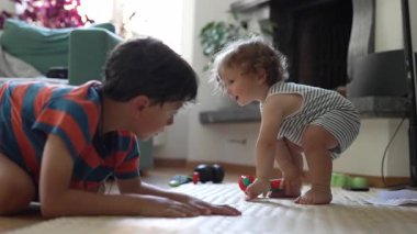 Oturma odasındaki küçük çocukla büyük kardeş arasında neşeli bir etkileşim, yerde sevgi dolu bir kucaklaşma yaşarlarken samimi bir bağ ve denge anı..