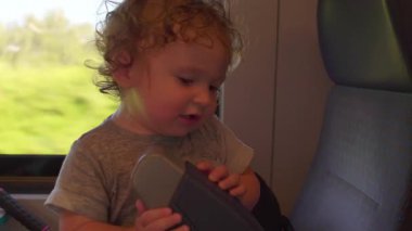 Kıvırcık saçlı çocuk, iki eliyle dizlik tutarak tren koltuğunun yanında duruyor. İç mekanı inceler ve keşfederken aşağı bakıyor.