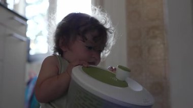 Bebek mutfakta oturmuş salata çeviricisiyle oynuyor. İki elinde yeşil plastik kapları inceliyor.