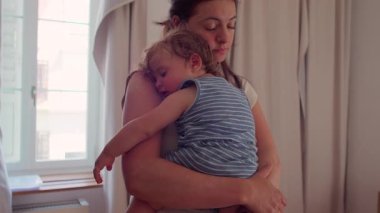 Anne pencerenin önünde duruyor uyuyan bebeği göğsüne yaslıyor, doğal ışık perdeleri ve camdan görülebilen bir bina.