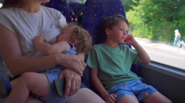 Bebek annelerin kucağında emzirirken büyük kardeş otobüs penceresinden dışarı bakar, gün ışığı her iki çocuğu da huzur içinde sürerken aydınlatır..
