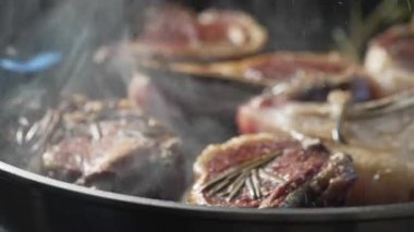 Kuzu pirzolası tavada pişiyor, biberiye ve hafif dumanla, yüzeyde altın kabuk gelişiyor, yağ ve kavrulmuş kenarlar parlıyor.