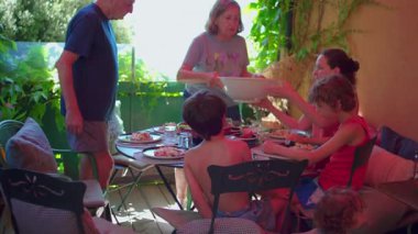 Aile, gölgeli bir verandada gündelik yaz yemeği için toplanıyor. Nesiller boyu masada oturup dışarıda yemek ve sohbet ediyorlar..
