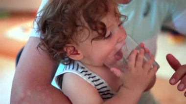 Yetişkin kollarında tutulan çocuk, boş cam bardaktan su içiyormuş gibi yapıyor, gözleri kapalı ve dudakları hassas bir anda aile evinde oynuyormuş gibi.