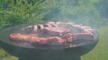 Güneşli bir günde kalın dumanlı, sosisli, pirzola ve tavuk ızgaralı geniş ölçekli barbekü, yeşil arka bahçede et cızırdıyor.