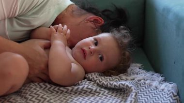 Anne bebeği yumuşak kanepede kucaklar çocuk kameraya sakince bakar, doğal aydınlatılmış oturma odasındaki battaniyede hassas bir anı paylaşır.