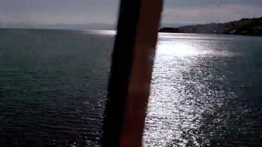 Güneş ışığı sakin göl yüzeyine yansıyor. Sahil kasabasının silueti ve sakin bir yaz öğleden sonrası boyunca ön planda bulanık ağaç dalları.