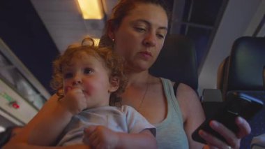 Kadın ve bebek trene yakın otururlar, kıvırcık saçlı bebek annesinin kucağına sakince oturur telefona bakarken, yumuşak odaklı arka plan.