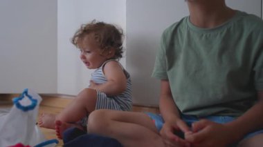 Küçük çocuk, ahşap evde, rahat bir evde, ahşap zeminde, dağınık oyuncaklar ve ev eşyalarıyla çevrili bir şekilde otururken utangaç bir şekilde gülümsüyor..