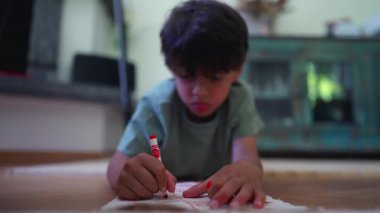 Evdeki düşünceli ifadeye odaklanmış, kırmızı kalemle kağıdı boyarken dilini hafifçe dışarı çıkaran genç bir çocuk.