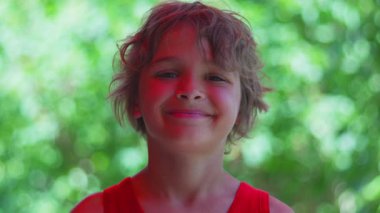 Çocuk kameraya doğru tüm yüzüyle gülümsüyor, yaz yeşillikleri tarafından aydınlatılıyor, kendinden emin ve kaygısız ifade doğal ortamda yakalanıyor.