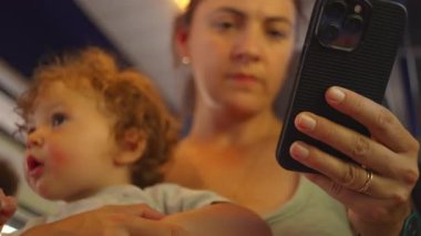 Kıvırcık saçlı bebek, hareket halindeki trende annesinin kollarında uzanır. Akıllı telefonları kontrol ederken gözlerini diker..