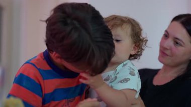 Bebek, kendinden büyük bir çocukla etkileşime girerken bir yandan da bir kadın tarafından tutuluyor. Günlük aile ortamında sevgi dolu bir kardeş ilişkisi.