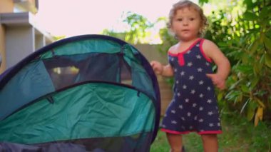 Bebek, güneşli arka bahçede oyun çadırının yanında yürür. Gözleri açık, tetikte olur. Anlık merak anında çerçeveden çıkar.