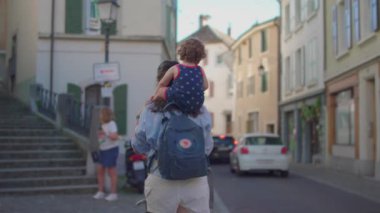 Anne, Avrupa 'nın eski şehrinde, renkli cepheleri, park halindeki arabaları ve arka plandaki yaya aktiviteleriyle kaldırım taşı caddesinde yürümeye devam ediyor..