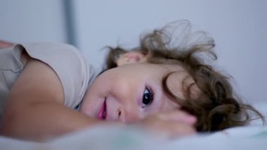 Bebek yatakta yatarken başını kaldırıyor, yumuşak ifade ve kıvırcık saçlarla kameraya bakıyor, kapalı alanda erken farkındalık anı ve nazik temas anı.