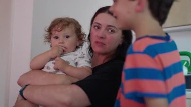 Anne kucağında bebek ve yanaktan nazikçe öpüşürken büyük çocuk yanında duruyor. Evin içinde özgün anne şefkati ve aile şefkati sahnesi.