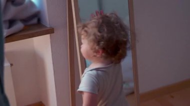 Küçük çocuk aynaya sarılıp kameraya doğru yürümeden önce arkasını dönüyor, kapalı alanda hareket, denge ve erken keşifler gösteriyor.