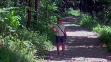 Kıdemli kadın ormanda tek başına yürüyor, elleri rahat, güneş gözlüğü takıyor, açık yaz ormanlarında yalnız başına doğa yürüyüşü yaparken