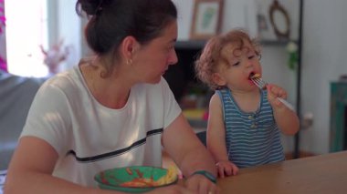 Küçük çocuk çatalla makarna yerken anne izliyor, her ikisi de günlük ev ortamında, parlak doğal ışıkla yemek masasında oturuyor.