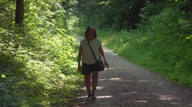 Yaşlı bir kadın, yemyeşil ormandaki çakıllı patikada tek başına yürüyor güneşli yaz sabahında, ağaçlar ve bitkilerle çevrili, huzur içinde tek başına yürüyüş yapıyor.