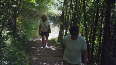 Yaşlı çift orman yolunda yürüyor, adam güneş gözlüğü takıyor ve şapka takıyor. Bu arada kadın yaz boyunca ağaçlarla kaplı patikayı takip ediyor.