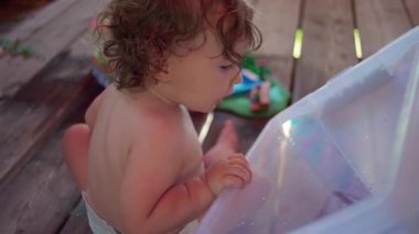 Yeni yürümeye başlayan çocuk terasta oturup büyük plastik kapları inceliyor, ılık yaz gününde su oyunu sırasında merakla içeriyi inceliyor.