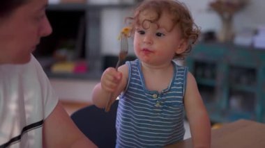 Yemek masasında çatal, dağınık yüz ve odaklanmış ifade ile makarnayı bağımsız olarak yiyen çocuk sıcak aile mutfağında ebeveynin yanında otururken