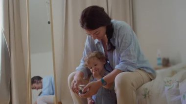 Annem bebeğe yabanmersini konteynırını tutmasında yardım ederken arka planda doğal ışık ve ayna yansıması olan sıcak yatak odasında rahat bir yatak odasında oturuyor.