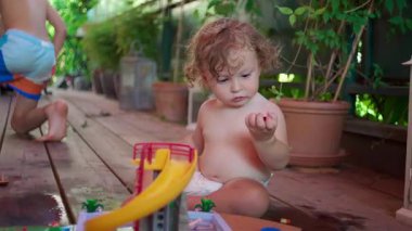 Küçük çocuk gölgeli veranda zemininde sürünerek plastik oyuncak kaydırağına uzanır, tamamen oyun zamanı aktivitelerine dalmış, renkli düzenekler ve arka planda yapraklı yeşil bitkiler vardır.