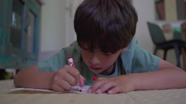 Yerde kırmızı kalemle çizim yapan genç bir çocuk, odaklanmış ifade, mobilyalarla dolu rahat bir iç mekan ve yumuşak arka planda soyut duvar sanatı.