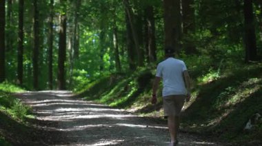 Kıdemli adam kıvrımlı patikada yürüyor. Parlak yeşil yapraklar ve dağınık güneş ışığı, beyaz gömlek ve şort giyiyor.
